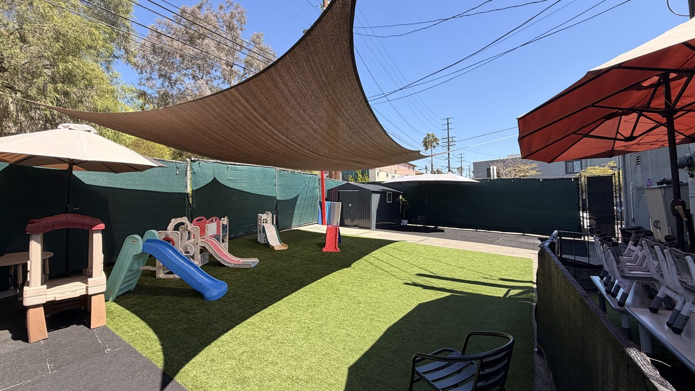 A shaded play yard at Sunshine Child Care Center, with a sail over artificial turf and toddler slides along the fence.